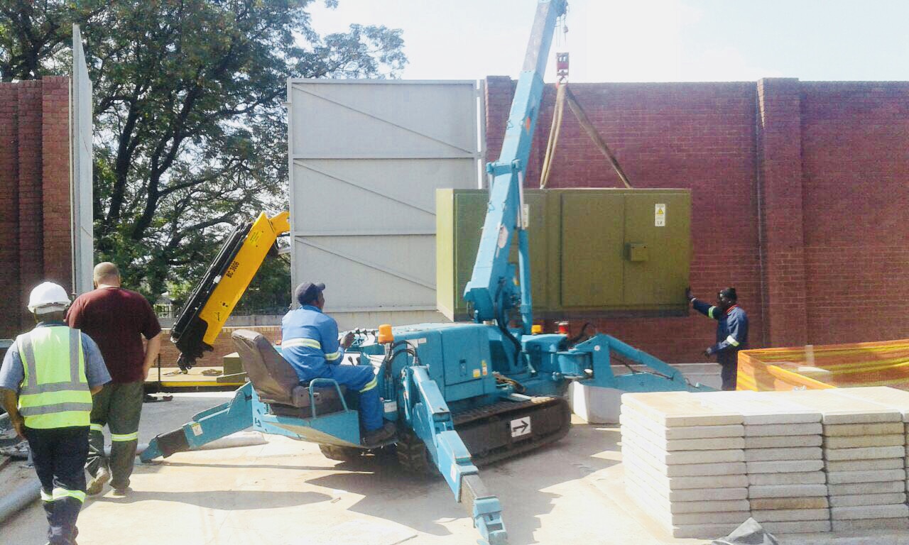 Teal spider crane on tracks lifting a container over a boundary wall